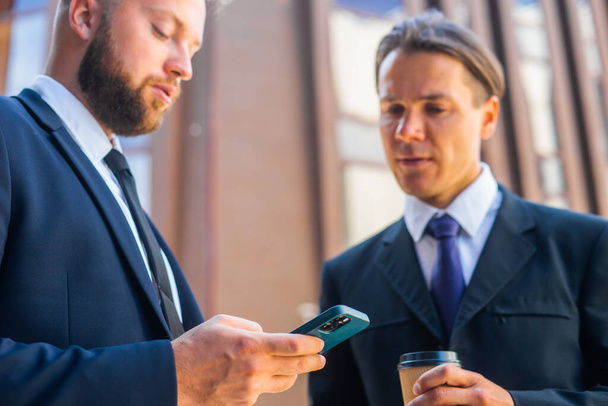 Confident businessman and his colleague are using smartphone in front of modern office building. Financial investors are talking outdoor. Banking and business. - Foto, imagen