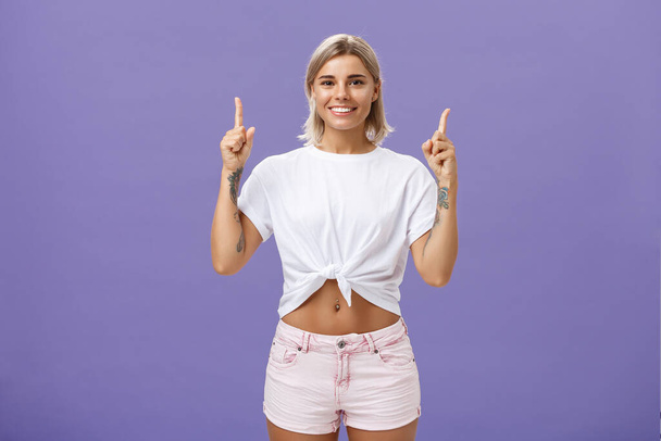 Time look upwards and move forward corporate ladder. Portrait of attractive ambitious and stylish young blonde woman with tattoos and pierced belly pointing up and smiling broadly over purple wall - Photo, Image