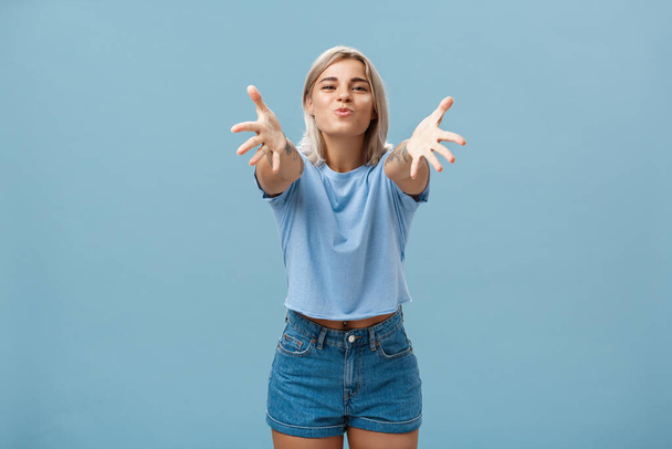 Come to mama. Portrait of friendly joyful and enthusiastic caucasian female student in trendy outfit pulling hands towads camera and folding lips to hug and give passionate kiss smiling over blue wall - Photo, Image