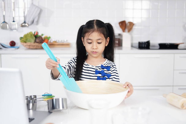 Asian little girl child is learning and enjoying for baking cookies bakery in a white bowl on wooden table in kitchen. Homemade pastry for bread. Family love and Homeschool. - Photo, Image