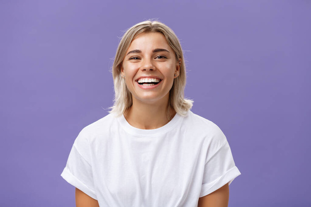 Κοντινό πλάνο της χαρούμενης γοητευτικής ξανθιάς με το ενθουσιασμένο και ευχαριστημένο χαμόγελο στέκεται σε λευκό μπλουζάκι πάνω από μωβ φόντο ξοδεύοντας χρόνο σε φοβερή διασκεδαστική παρέα - Φωτογραφία, εικόνα