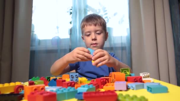 Kaunis poika leikkii kotona rakennuspalikoilla. Söpö hymyilevä poika leikkii lego-rakentajan kanssa, jolla on paljon värikkäitä muovisia lohkoja huoneessa, rakentamassa kaupunkia. Esikoululuokat. - Materiaali, video