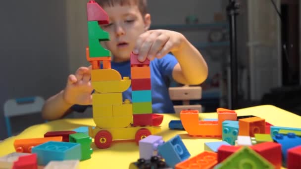 Güzel bir çocuk evde yapı taşlarıyla oynuyor. Şirin, gülümseyen bir çocuk, bir sürü renkli plastik bloğu olan bir lego inşaatçısıyla oynuyor ve bir şehir inşa ediyor. Anaokulu sınıfları. - Video, Çekim