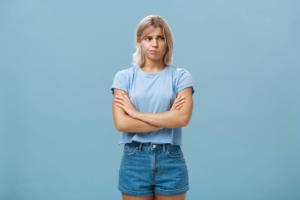 Uncertain troubled and perplexed attractive blond woman with tanned skin holding hands crossed on chest pouting and frowning looking right with worried unsure and doubtful look over blue wall - Photo, Image