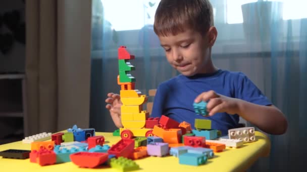 Krásný chlapec si hraje doma se stavebními kameny. Roztomilý usměvavý chlapec si hraje s konstruktérem lega se spoustou barevných plastových bloků v místnosti, buduje město. Předškolní třídy. - Záběry, video