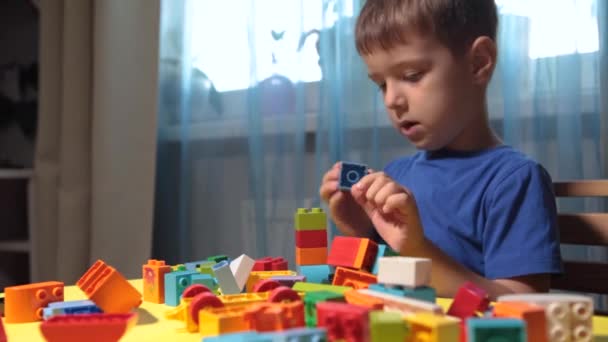 Un beau garçon joue à la maison avec des blocs de construction. Un garçon souriant mignon joue avec un constructeur de lego avec beaucoup de blocs en plastique colorés dans la pièce, la construction d'une ville. Cours préscolaires. - Séquence, vidéo