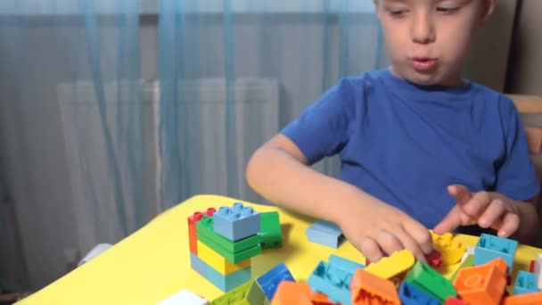 Egy gyönyörű fiú játszik otthon építőkockákkal. Egy aranyos mosolygós fiú játszik egy lego konstruktorral, sok színes műanyag tömbbel a szobában, várost építve. Óvodai osztályok. - Felvétel, videó