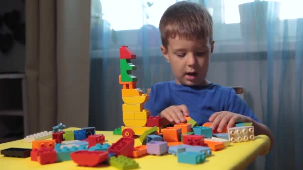 Kaunis poika leikkii kotona rakennuspalikoilla. Söpö hymyilevä poika leikkii lego-rakentajan kanssa, jolla on paljon värikkäitä muovisia lohkoja huoneessa, rakentamassa kaupunkia. Esikoululuokat. - Materiaali, video
