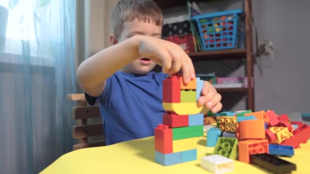 Un beau garçon joue à la maison avec des blocs de construction. Un garçon souriant mignon joue avec un constructeur de lego avec beaucoup de blocs en plastique colorés dans la pièce, la construction d'une ville. Cours préscolaires. - Séquence, vidéo