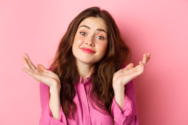 Close up portrait of clueless cute woman, shrugging shoulders and smiling with unaware face expression, standing over pink background - Photo, Image