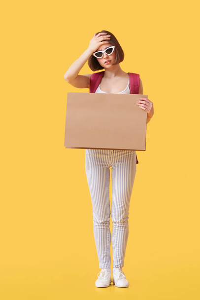 Upset young woman with cardboard hitchhiking on color background - Foto, afbeelding