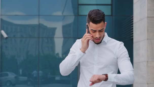 Portrait successful serious arabic hispanic business man spaniard boss leader wears white formal shirt stands outdoors speaks mobile phone cellphone answers call looks at wrist watch checks late time - Footage, Video