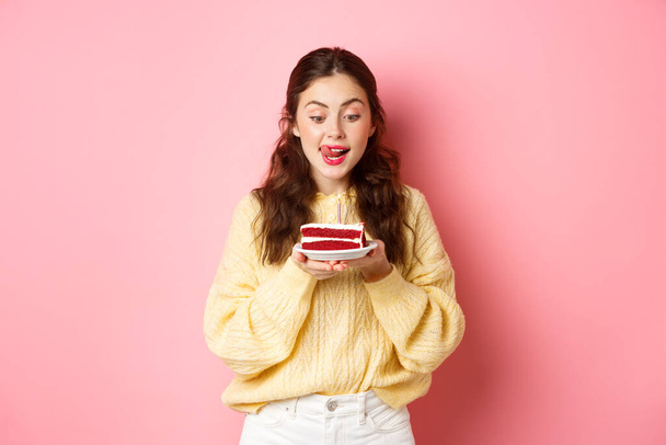 Feestdagen en feestdagen. Happy birthday girl likken haar lippen en kijk met verleid gezicht op b-day cake, wil bijten, staande tegen roze achtergrond - Foto, afbeelding