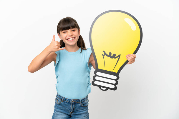 Little caucasian kid isolated on white background holding a bulb icon and doing phone gesture - Foto, imagen