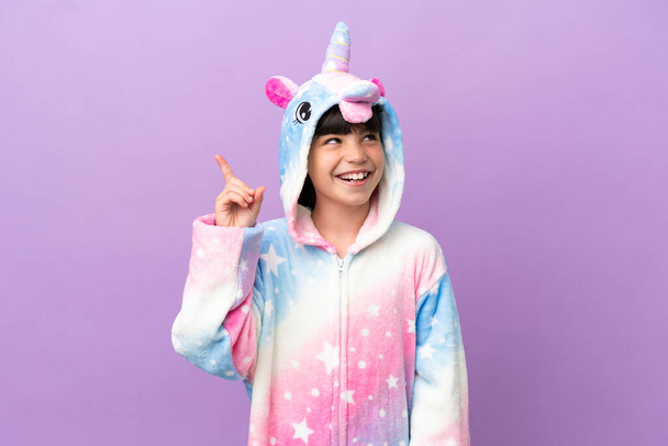 Kleines Kind im Einhorn-Pyjama isoliert auf violettem Hintergrund mit der Absicht, die Lösung zu erkennen, während es den Finger hebt - Foto, Bild