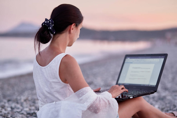 Mladá brunetka s laptopem na lehátku na pláži. Freelancer, práce na dálku, webinář, učení koncepce, chatování s přáteli. Nový normální koncept. - Fotografie, Obrázek