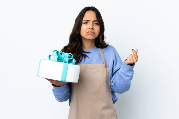 Pastry chef holding a big cake over isolated white background making money gesture but is ruined - Photo, Image