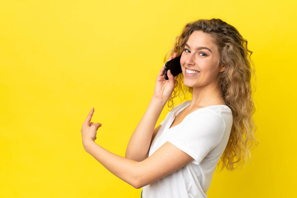 Mujer rubia joven usando teléfono móvil aislado sobre fondo amarillo apuntando hacia atrás - Foto, imagen
