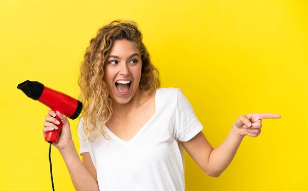 Jovem loira segurando um secador de cabelo isolado no fundo amarelo apontando o dedo para o lado e apresentando um produto - Foto, Imagem