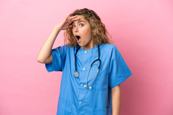 Junge Chirurgin Ärztin isoliert auf rosa Hintergrund macht überraschende Geste, während sie zur Seite schaut - Foto, Bild