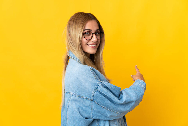 Young blonde woman isolated on yellow background pointing back - Zdjęcie, obraz
