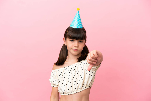 Klein blank kind met verjaardagshoed geïsoleerd op roze achtergrond met duim naar beneden met negatieve expressie - Foto, afbeelding