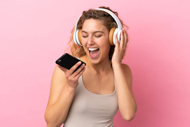 Νεαρή ξανθιά γυναίκα απομονωμένη σε ροζ φόντο ακούγοντας μουσική με κινητό και τραγουδώντας - Φωτογραφία, εικόνα