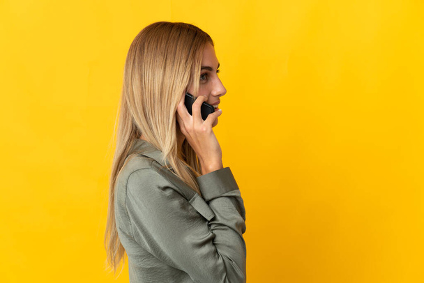 Young blonde woman isolated on yellow background keeping a conversation with the mobile phone with someone - Fotó, kép