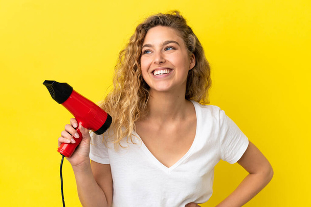Young blonde woman holding a hairdryer isolated on yellow background posing with arms at hip and smiling - Fotó, kép