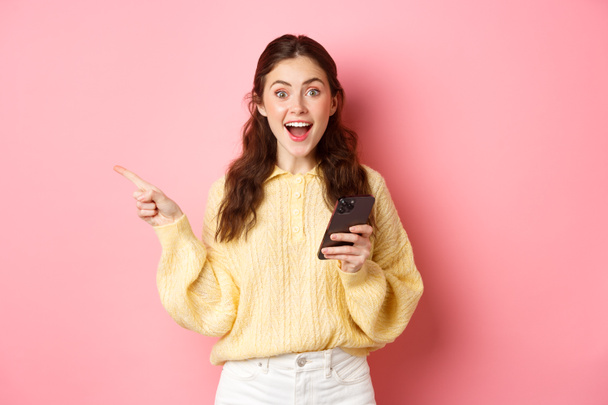 Izgalmas fiatal nő kezében mobiltelefon, ujjal mutogatott félre a bal copyspace, bemutató hirdetés, talált érdekes promo online okostelefon, szemben rózsaszín háttér - Fotó, kép