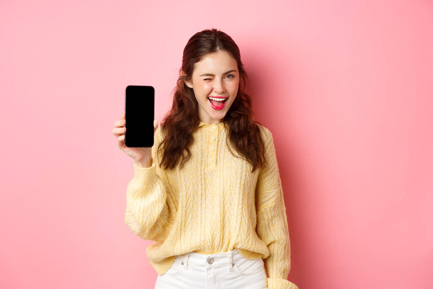 Wizerunek bezczelnej atrakcyjnej dziewczyny, pokazującej ekran smartfona, mrugającej i uśmiechniętej, polecającej telefon komórkowy, stojącej na różowym tle - Zdjęcie, obraz