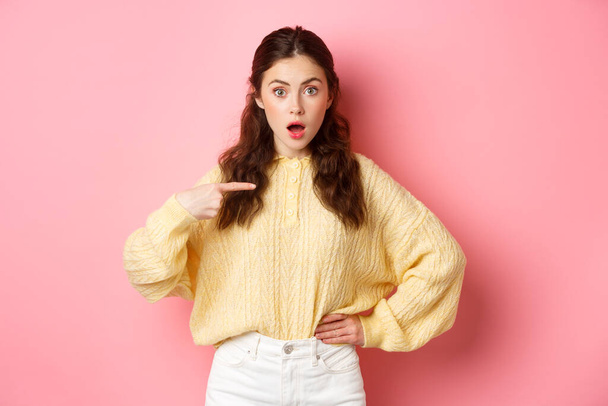 Young surprised woman gasping and pointing at herself with disbelief, being chosen or named, standing against pink background - Foto, afbeelding