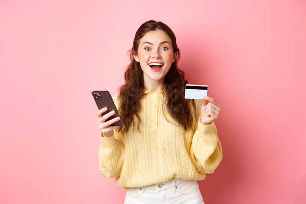 Технологии и интернет-магазины. Возбужденная девушка делает заказ, оплата онлайн с пластиковой кредитной картой, держа мобильный телефон и улыбаясь в камеру, розовый фон - Фото, изображение