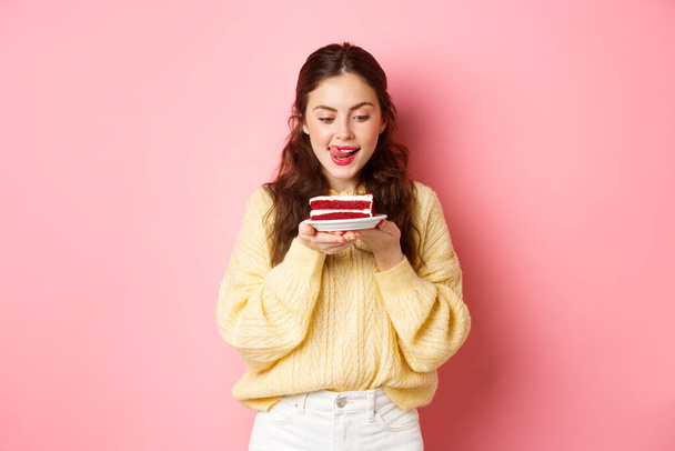 Young slim girl lick her lips from temptation, looking at delicious piece of cake with yearning to bite it, standing with dessert against pink background - Fotografie, Obrázek