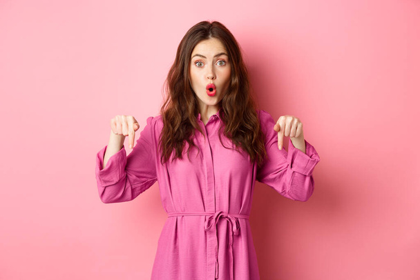 Stylish caucasian woman saying wow, pointing fingers down at impressive banner news, showing advertisement, staring amazed, standing against pink background - Fotó, kép