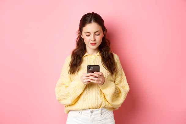 Technologie. Junge lächelnde Frau plaudert in sozialen Medien, liest Nachricht auf Smartphone, steht vor rosa Hintergrund - Foto, Bild
