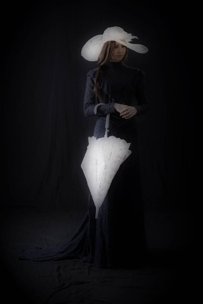 A girl in an old dress and hat stands on a dark background and is sad - Zdjęcie, obraz