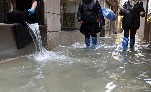 εργάτης αδειάζει το νερό από το κατάστημά της με έναν κουβά και άλλους ανθρώπους με αδιάβροχους βάτες καθώς περπατούν στο δρόμο πλημμυρίζοντας εντελώς από την παλίρροια στη Βενετία Ιταλία - Φωτογραφία, εικόνα