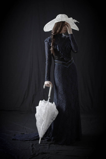 A girl in an old dress and hat stands on a dark background and is sad - Fotoğraf, Görsel