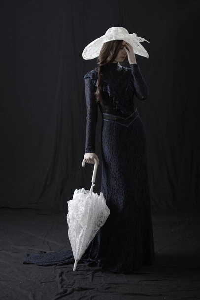 A girl in an old dress and hat stands on a dark background and is sad - Φωτογραφία, εικόνα