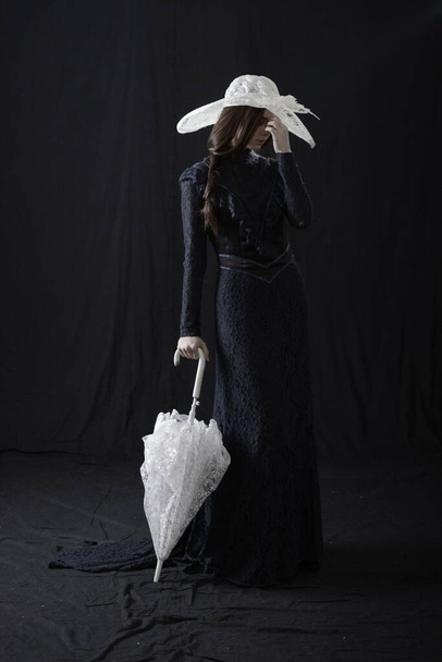 A girl in an old dress and hat stands on a dark background and is sad - Foto, Imagen