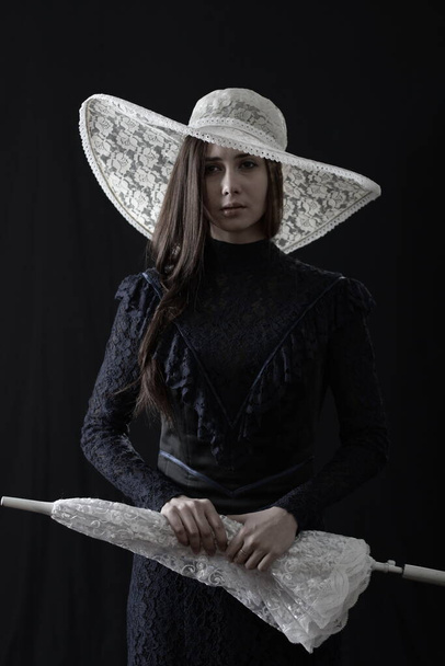 A girl in an old dress and hat stands on a dark background and is sad - Photo, image