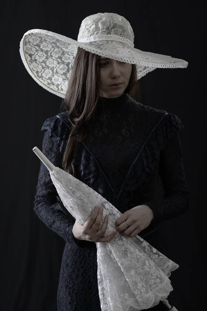 A girl in an old dress and hat stands on a dark background and is sad - 写真・画像