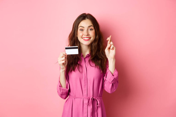 Image d'une jeune femme pleine d'espoir montrant une carte de crédit en plastique et des doigts croisés pour la bonne chance, faisant vœu et souriant, debout sur fond rose - Photo, image