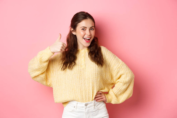 Portrait of beautiful brunette female model showing thumbs up, like your idea, give support, approve or praise something good, standing against pink background - Photo, Image