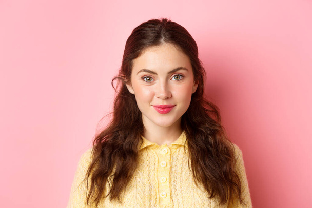 Close up of young 20s female model with cute feminine make up, smiling and looking hopeful at camera, standing against pink background - Photo, Image