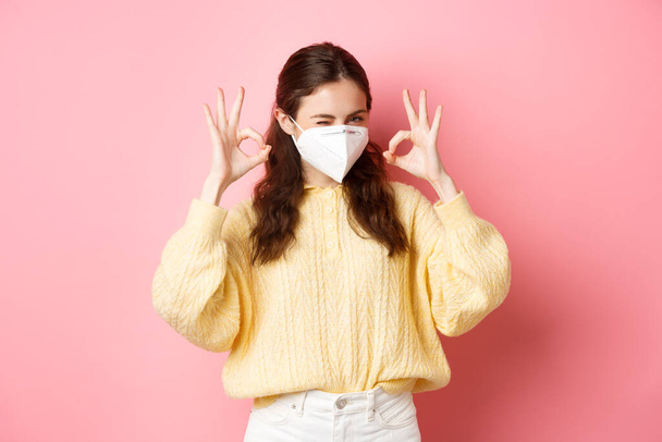 Covid-19, lockdown and pandemic concept. Young smiling girl assure you, winking and showing okay sign, alright or approve gesture, wearing medical respirator, standing over pink background - 写真・画像