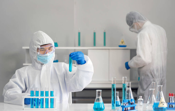 Epidemiological researcher in virus protective clothing look at  blue liquid chemicals in test tube. Coronavirus disease 2019 (COVID-19) testing process in a laboratory. - Photo, Image