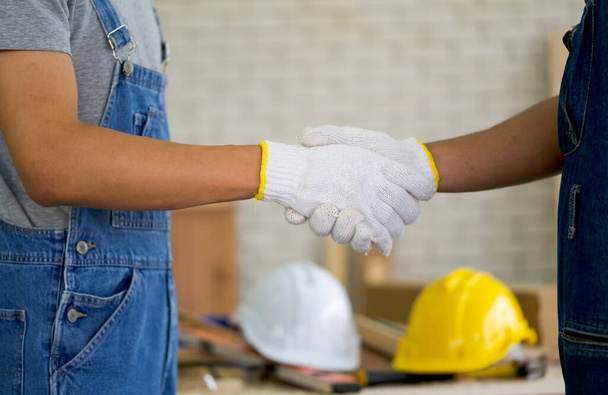 Carpenters with protective glove shaking hands after finish the job. A white and yellow construction helmet was placed on the table in the background. - Foto, Bild