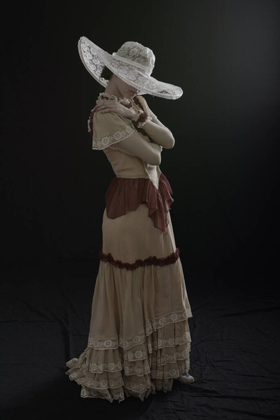 A girl in an old dress and hat stands on a dark background and is sad - Foto, imagen